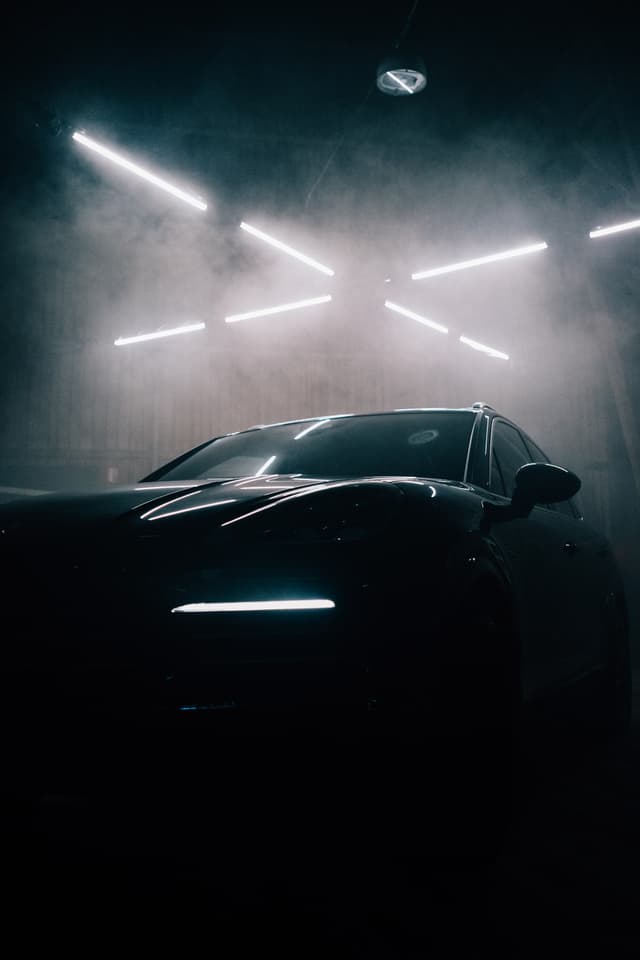 Dark-colored car in a painting booth with paint mist above it and overhead lights